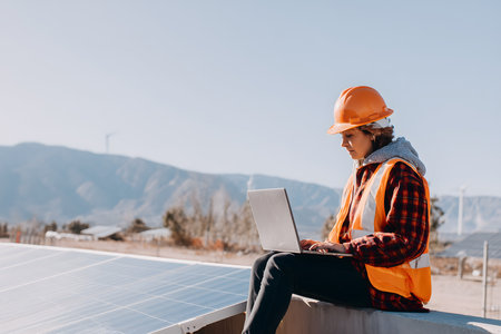 Female engineer wearing safety vest and helmet using laptop while sitting next to solar panels at power plantの素材