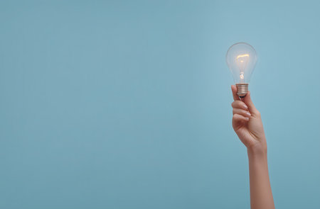 Hand holding a glowing light bulb against a vibrant blue backdrop, representing new ideas, innovation, and problem-solvingの素材