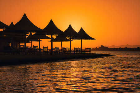 Beach umbrellas and tables creating a silhouette against a beautiful orange sunset on a tropical island, evoking a sense of tranquility and relaxationの素材