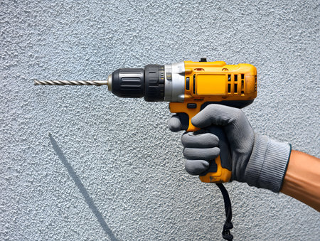 Hand wearing protective glove holding cordless electric drill preparing to make hole in concrete wallの素材