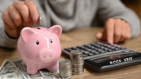 Accountant inserting coin in piggy bank while calculating home budget with calculator and dollar banknotes on wooden deskの素材