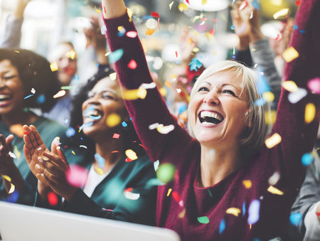 Diverse group of happy colleagues cheering and clapping with raised arms, celebrating achievement and joy with confettiの素材
