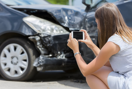 Woman crouching, capturing impact damage to a dark vehicle with a smartphone for insurance purposes after a road collisionの素材