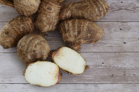 Taro corms with one sliced to reveal starchy white interior on rustic light wooden surface, overhead...