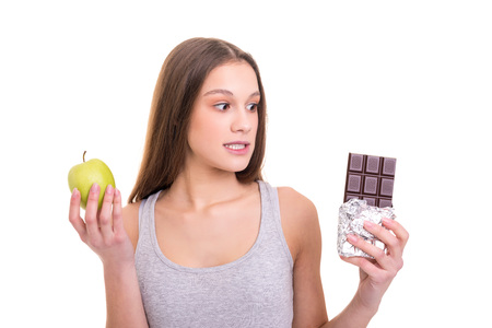 Thoughtful young blonde woman hesitating between a fruit and chocolate, isolated over white backgroundの写真素材
