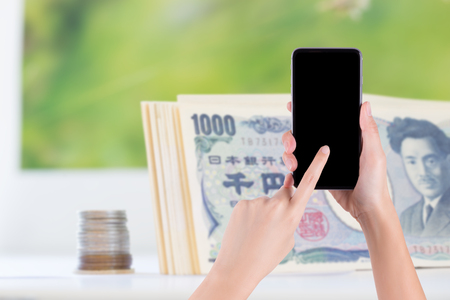 woman's hand holding smartphone and pointing on blank screen with japanese money on backgroundの写真素材