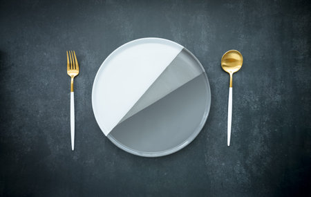 blank round white and grey ceramic dish with brass spoon and fork on dark blue background, food and...