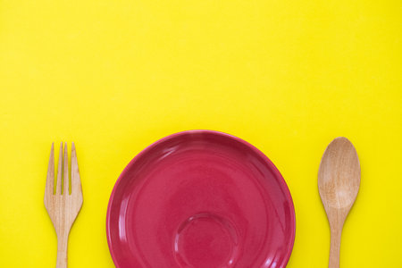 blank round red ceramic dish with wood spoon and fork on yellow background, food and drink, dining conceptの写真素材