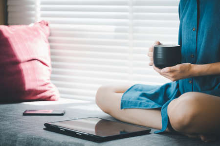 closeup casual dressed woman sitting on couch holding cup of coffee looking at tablet, work from home conceptの写真素材