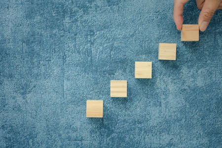 fingers choosing top wood cube arranged as stairs on blue background, business conceptの写真素材