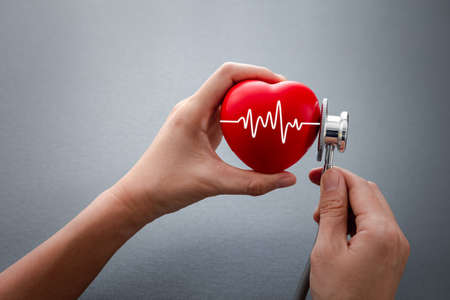 hands holding stethoscope checking on red rubber heart with cardiogram on dark grey background, health conceptの写真素材
