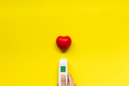 hand hold infrared thermometer and red heart on yellow background, healthcare and medical concept