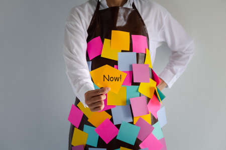 closeup woman wears apron with colorful sticky note paper holding a note with now word on one hand, concept and idea showing busyの写真素材