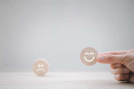 closeup woman hand picking happy emotion face on round wooden piece on table with sad on on background for satisfaction survey, mental health assessmentの写真素材