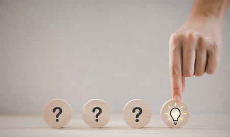closeup hand holding round wood piece with light bulb icon among question mark pieces, thinking and idea conceptの写真素材