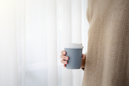closeup woman hand holding paper cup of hot coffee with semi transparent white curtain, morning lifestyleの写真素材