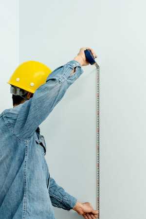 A construction worker wearing a yellow hard hat is measuring the wall with a tape measure. Home and...