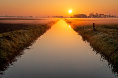 Atmospheric sunrise in the Netherlands with a beautiful orange glow in the air and water vapor over the fields and the canal and pondsの写真素材