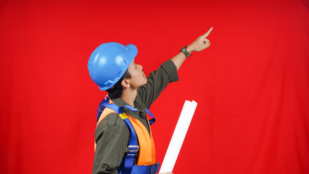 Asian worker man wear unifrom, helmet, safety harness and paper project pointing to blank space isolated red backgroundの写真素材