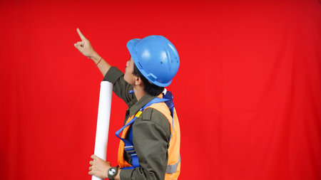 Asian worker man wear unifrom, helmet, safety harness and paper project pointing to left blank space isolated red backgroundの写真素材