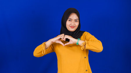 Portrait of Asian muslim woman smiling at camera and making love or heart sign gesture with her hand isolated blue backgroundの写真素材
