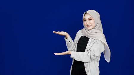 Asian hijab woman pointing left blank space isolated blue background