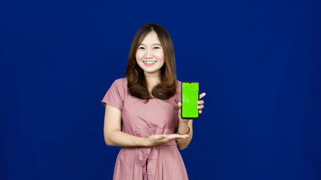beautiful asian woman pointing green screen in handphone isolated blue background