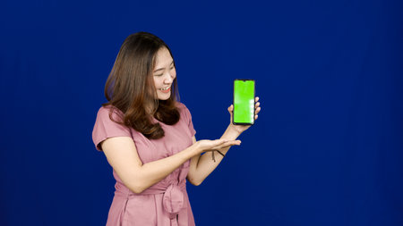 beautiful asian woman pointing green screen in handphone isolated blue backgroundの写真素材