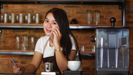 Young Asian woman barista wear apron talking and receive order from customer on cellphone at coffee shop. Concept of cafe store small business. female bartender writing note while listening clientの写真素材