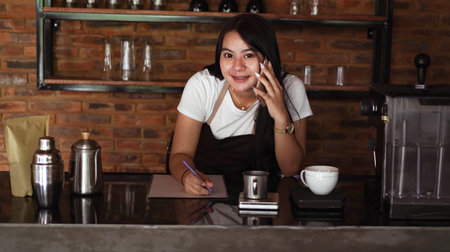 Young Asian woman barista wear apron talking and receive order from customer on cellphone at coffee shop. Concept of cafe store small business. female bartender writing note while listening clientの写真素材