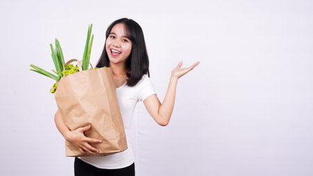 Beautiful Asian woman holding paper bag of fresh vegetables very happy pointing with hand and finger on isolated white backgroundの写真素材