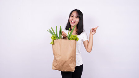 Beautiful Asian woman holding paper bag of fresh vegetables very happy pointing with hand and finger...