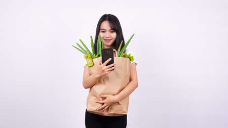 Asian woman with paper bag of fresh vegetables and holding a phone with isolated white backgroundの写真素材