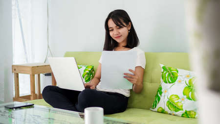 Young beautiful Asian woman working from home using laptop and sitting on chairの写真素材