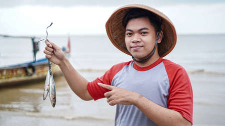 Happy young fisherman on the beach holding his catch fish and shows in front of his boatの写真素材