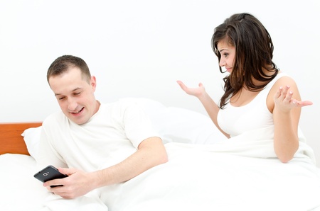 Young beautiful couple in bed, man is busy with his cell phone - woman upsetの写真素材