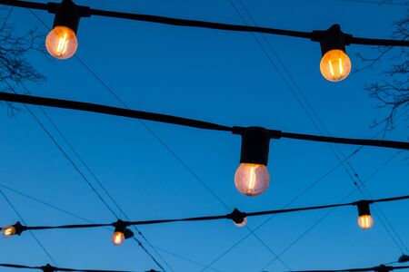 Light bulbs against the evening blue sky. Lanterns glowing with a pleasant warm yellow light.の写真素材