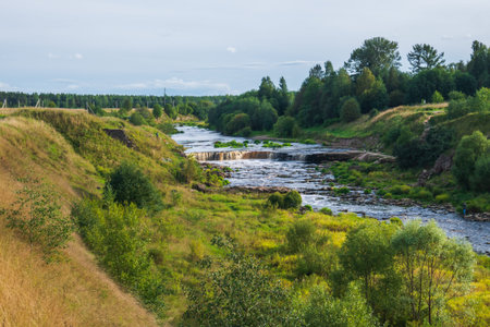 The coast of a small river, a large number of stones and greenery. Small house on the mountain.