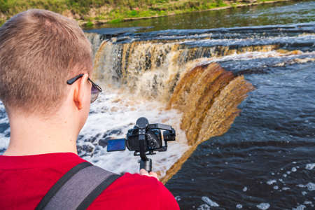 A male videographer walks on the water and films the current of the river. A photographer in a red T-shirt films as a stream of water rushes down.の写真素材