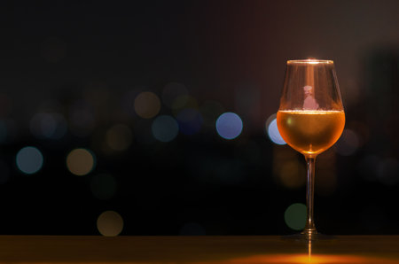 A glass of white wine on wooden table of rooftop bar with colorful bokeh of city light and space for text.の写真素材