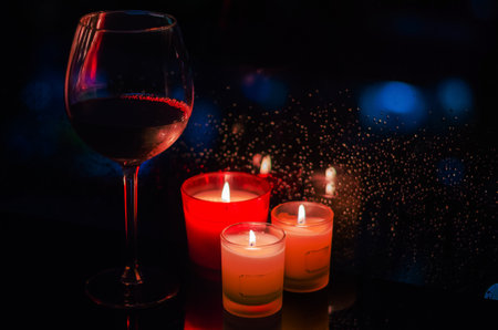 A glass of red wine with burning candles put beside window that have rain drop on dark background.の写真素材