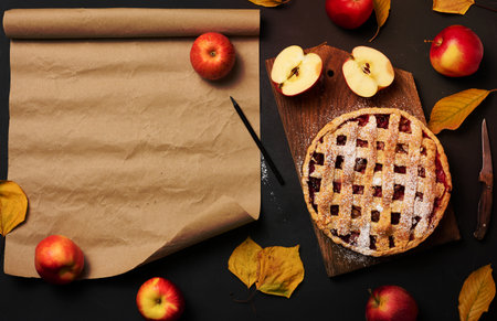 baked round traditional apple pie on a brown wooden board and fresh red apples. Nearby is a roll of brown paper for writing a recipe or menu, top viewの写真素材