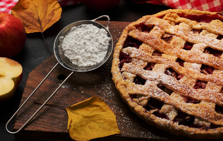 baked round traditional apple pie on a brown wooden board and fresh red apples. Next to it is an iron strainer with white powdered sugarの写真素材