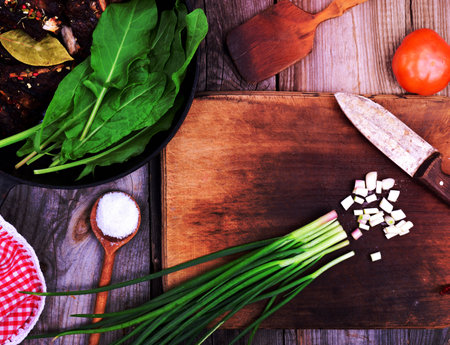 Chopped green onion on a wooden kitchen board, next to a frying pan with fried meatの写真素材