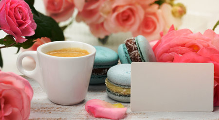 blank white business card and cup with espresso coffee and white ceramic cup with coffee and blue macaron on a white table, behind a bouquet of pink rosesの写真素材