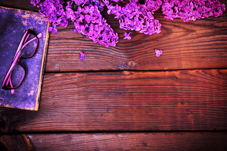 branch of a purple lilac and an old book with glasses on a brown wooden background, an empty spaceの写真素材