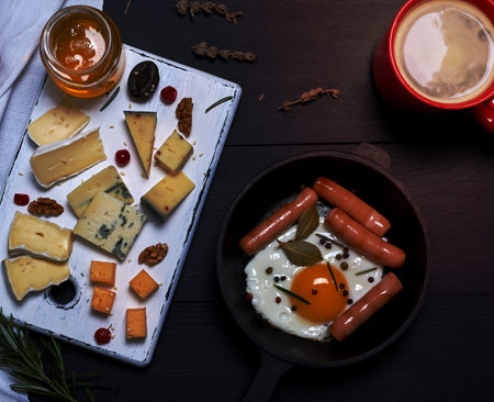 fried egg with sausages in a black round frying pan with a handle, a board with sliced different cheese and a cup of coffee, breakfast for one, top viewの写真素材