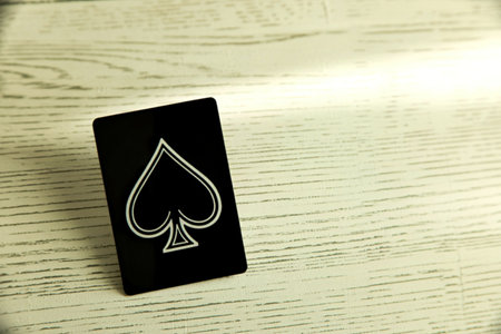 A close-up shot focuses on a black playing card featuring a prominent white spade symbol. The card lies diagonally on a light-colored wooden surface with visible grain.の写真素材
