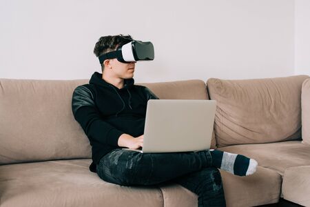 Man in black with glasses of virtual reality on couch. Young guy in VR headset is looking at interactive screen. Playing mobile game app on device. Home office VR.の写真素材