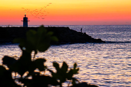 The silhouette of a man fishing on a rocky beach at sunset. Space for text.. Lighthouse and birds.の写真素材